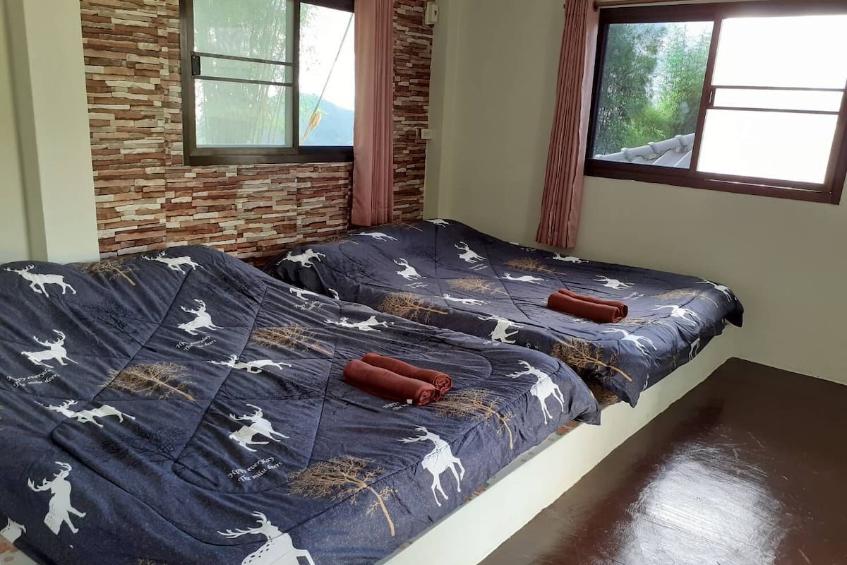 A room with two beds, each with a dark blue bedspread featuring white deer patterns and a slogan. There are two windows with pink curtains, one with a view of greenery outside.