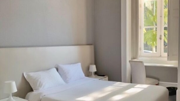 A bedroom with a large bed, a nightstand, a chair, and a window with a view of greenery. - WW Hostel & Suites (Coimbra)