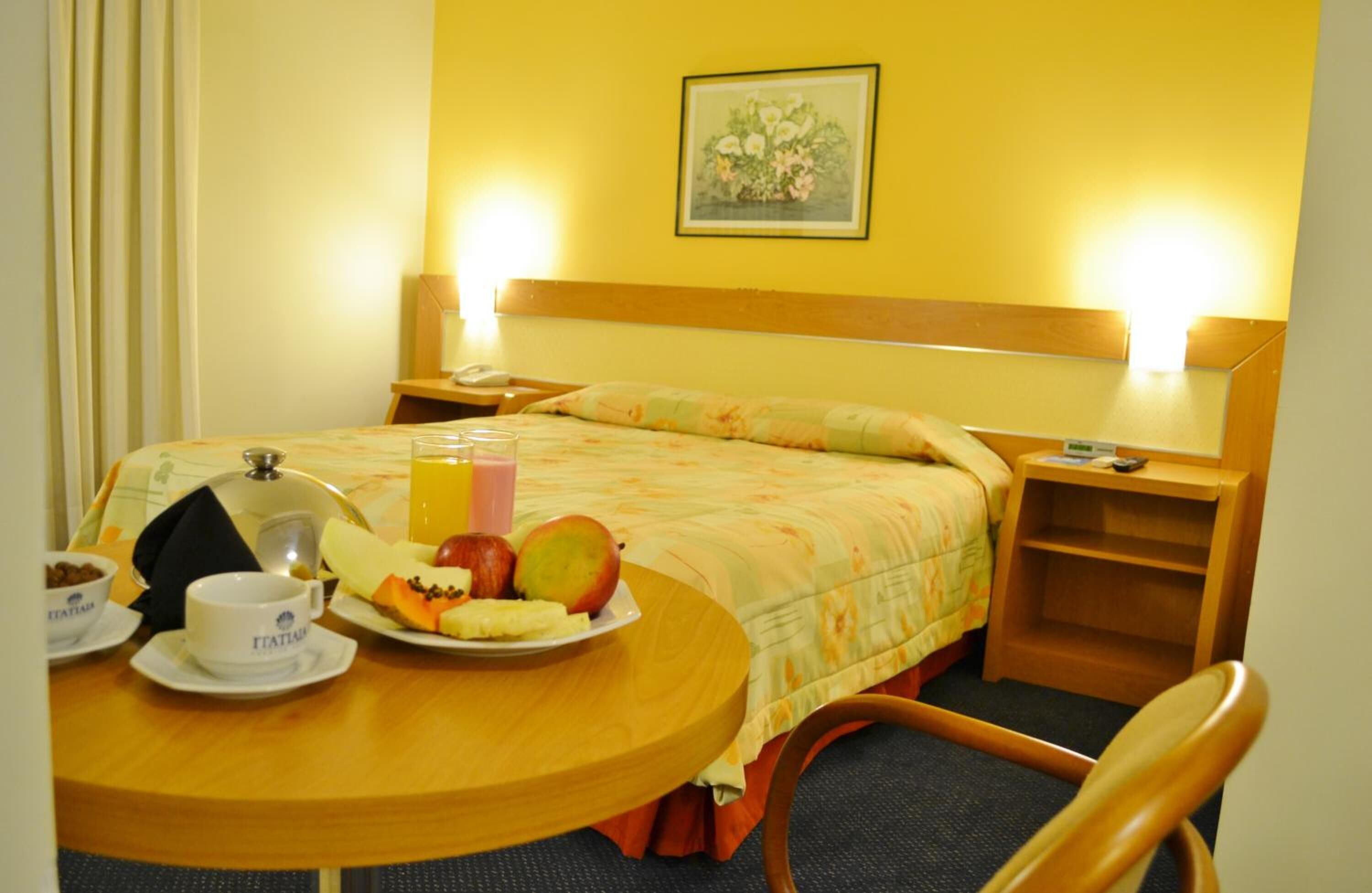 Una habitación de hotel con una cama, una mesita de noche con lámpara, un pequeño tocador de madera y una mesa de comedor con alimentos para el desayuno.