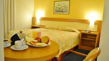 Una habitación de hotel con una cama, una mesita de noche con lámpara, un pequeño tocador de madera y una mesa de comedor con alimentos para el desayuno.