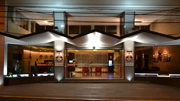 Una entrada de hotel moderna con fachada de vidrio, un mostrador de recepción y una escalera que lleva al piso superior.