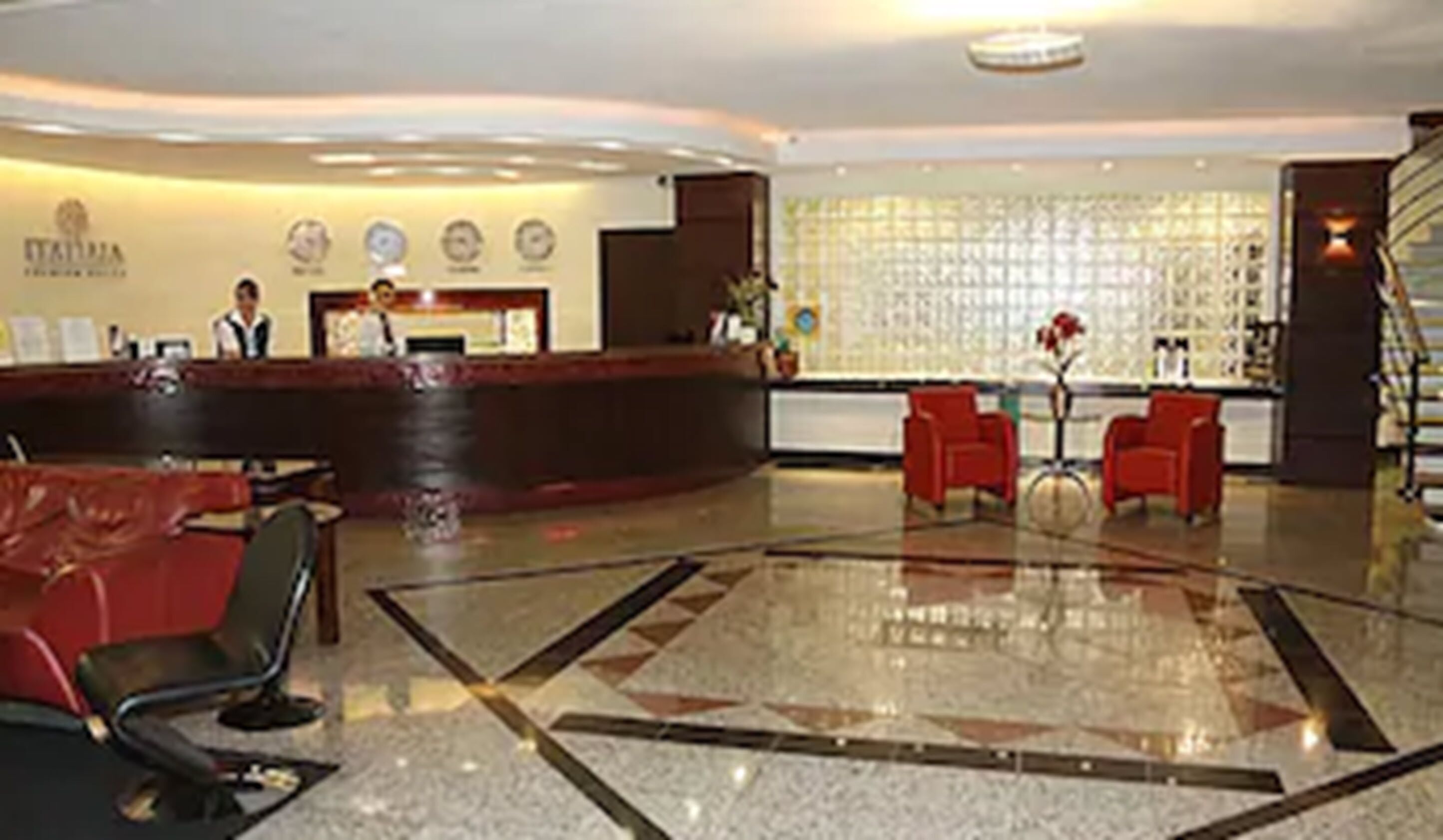Vestíbulo de hotel con mostrador de recepción, sillas rojas y piso estampado.