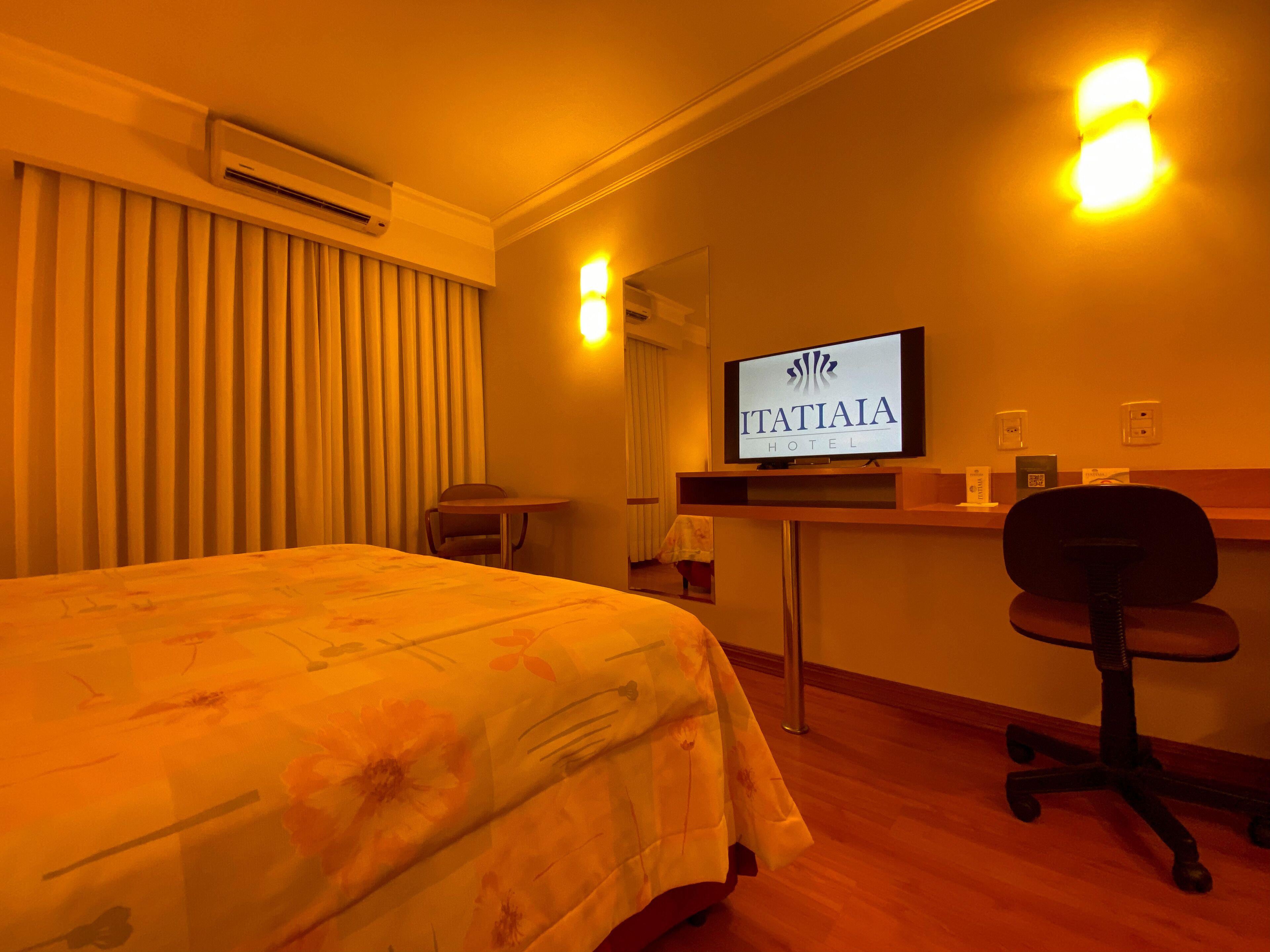 A hotel room with a bed, a desk, a chair, a television displaying the hotel name, and an air conditioning unit.
