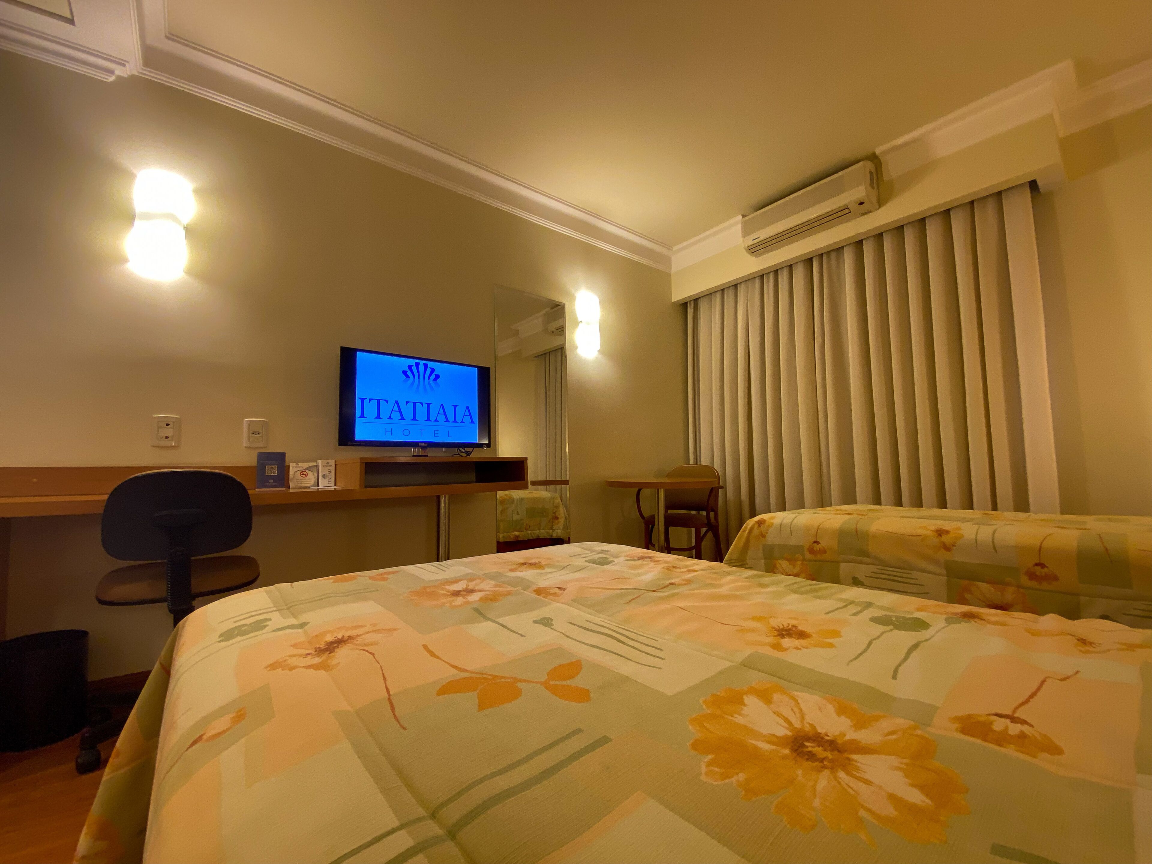 A hotel room with a bed, a desk, a chair, a television displaying the hotel name, and an air conditioning unit.