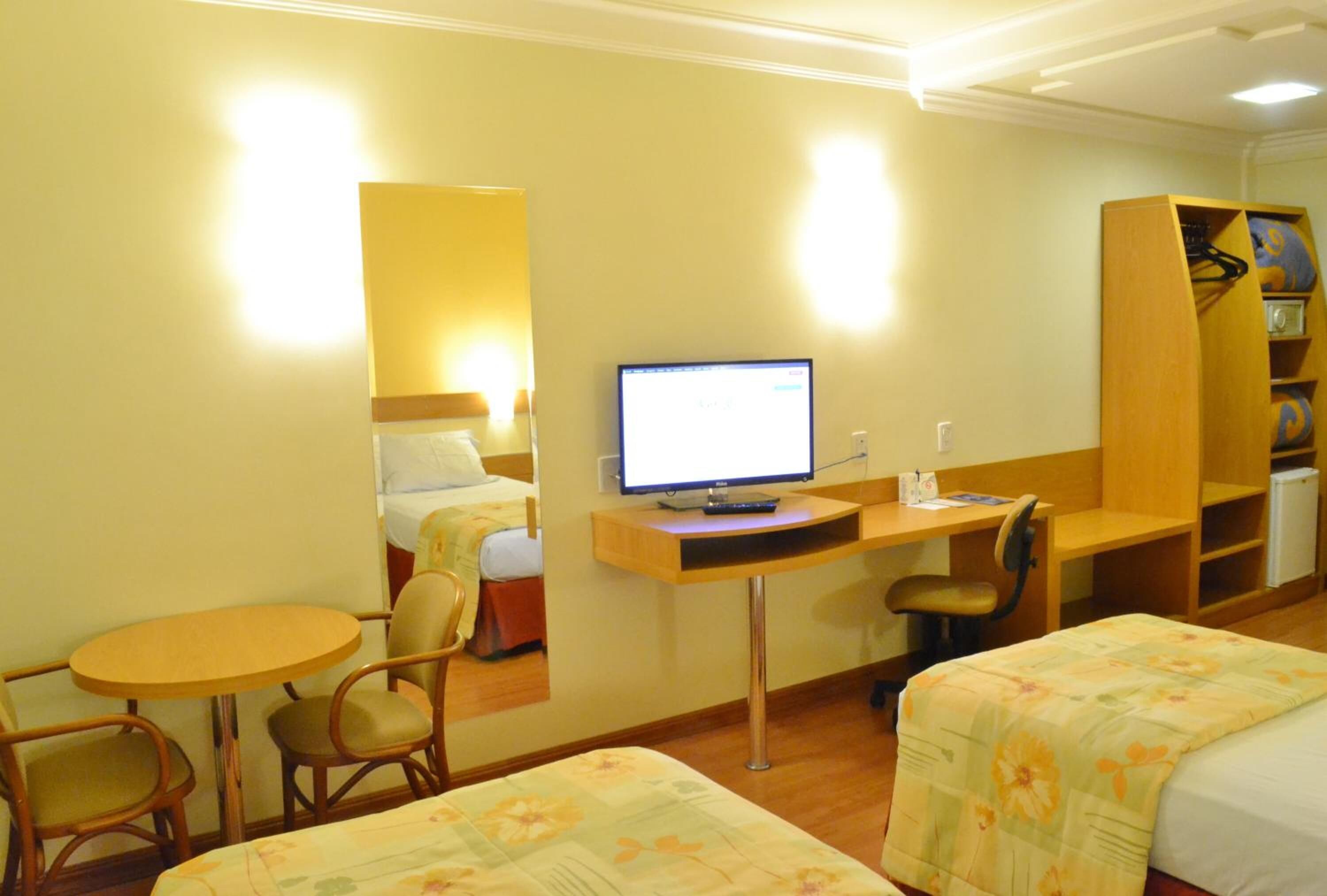 Una habitación de hotel con una cama, un escritorio con ordenador, una silla, una mesita y una estantería.
