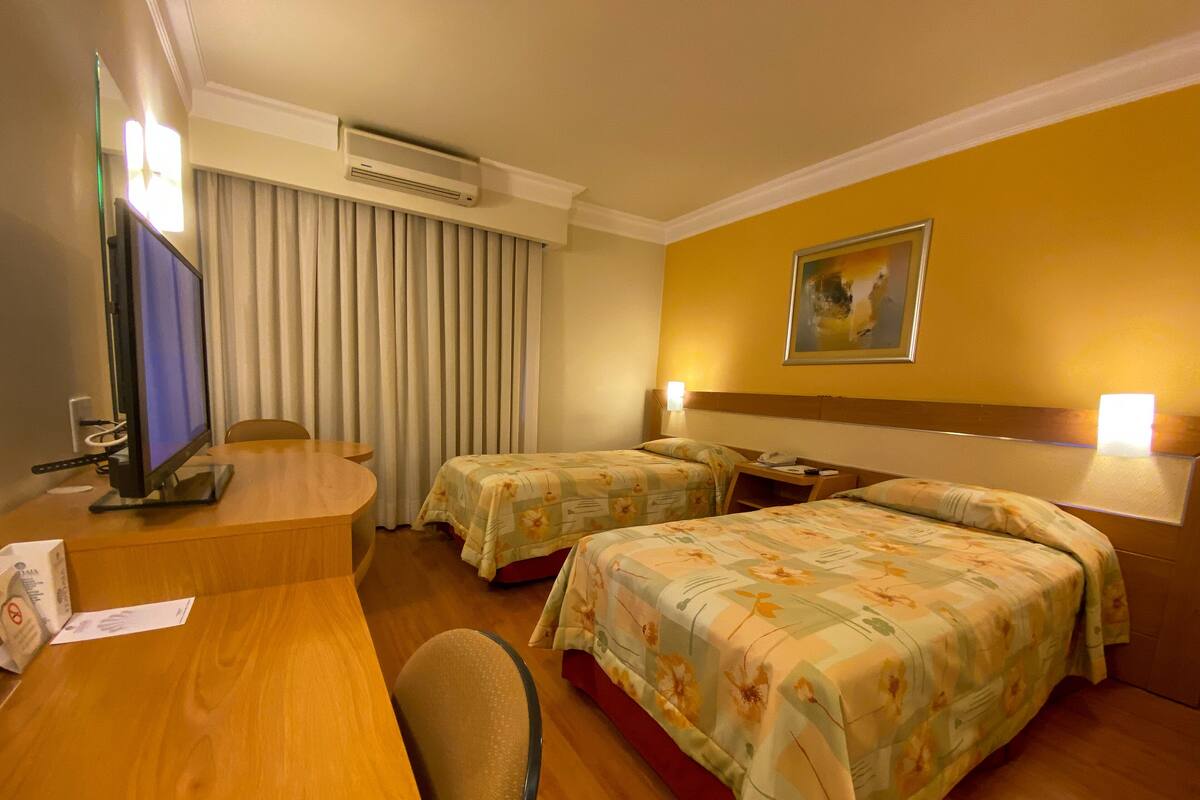 A hotel room with a desk, two beds, a TV, and an air conditioning unit.