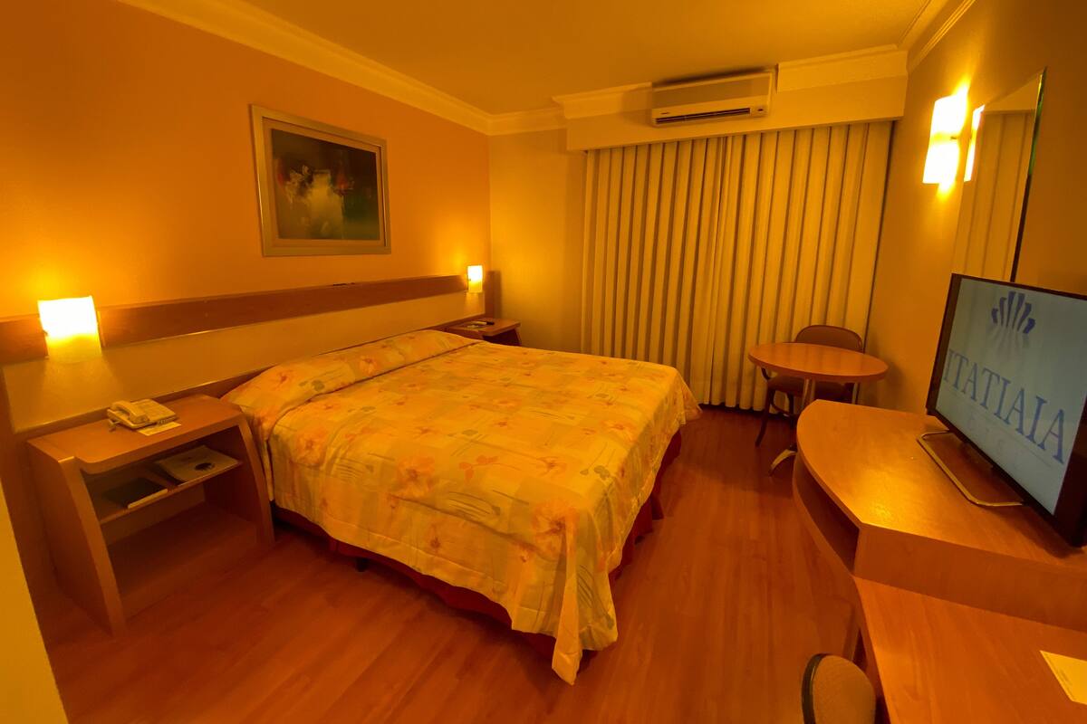 A hotel room with a bed, bedside table, TV, and a desk with a chair.