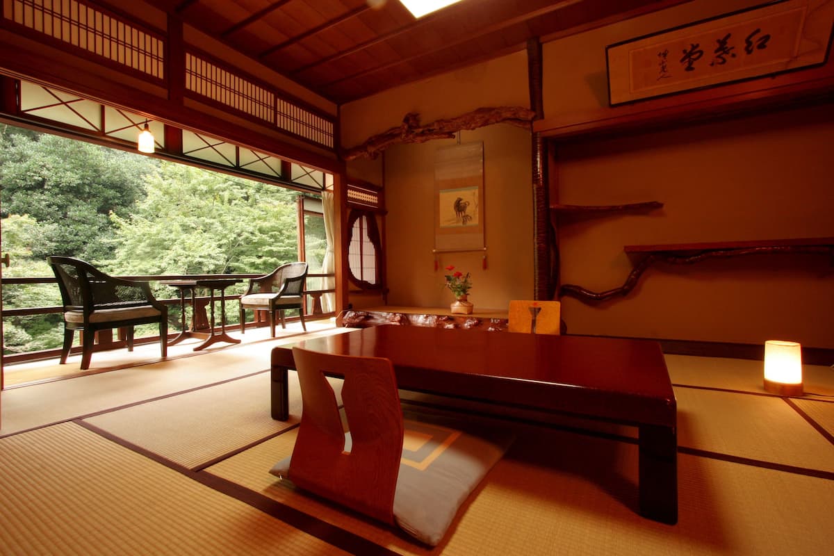 A traditional Japanese room with tatami flooring, a low table, and chairs. There is a view of greenery through the windows.