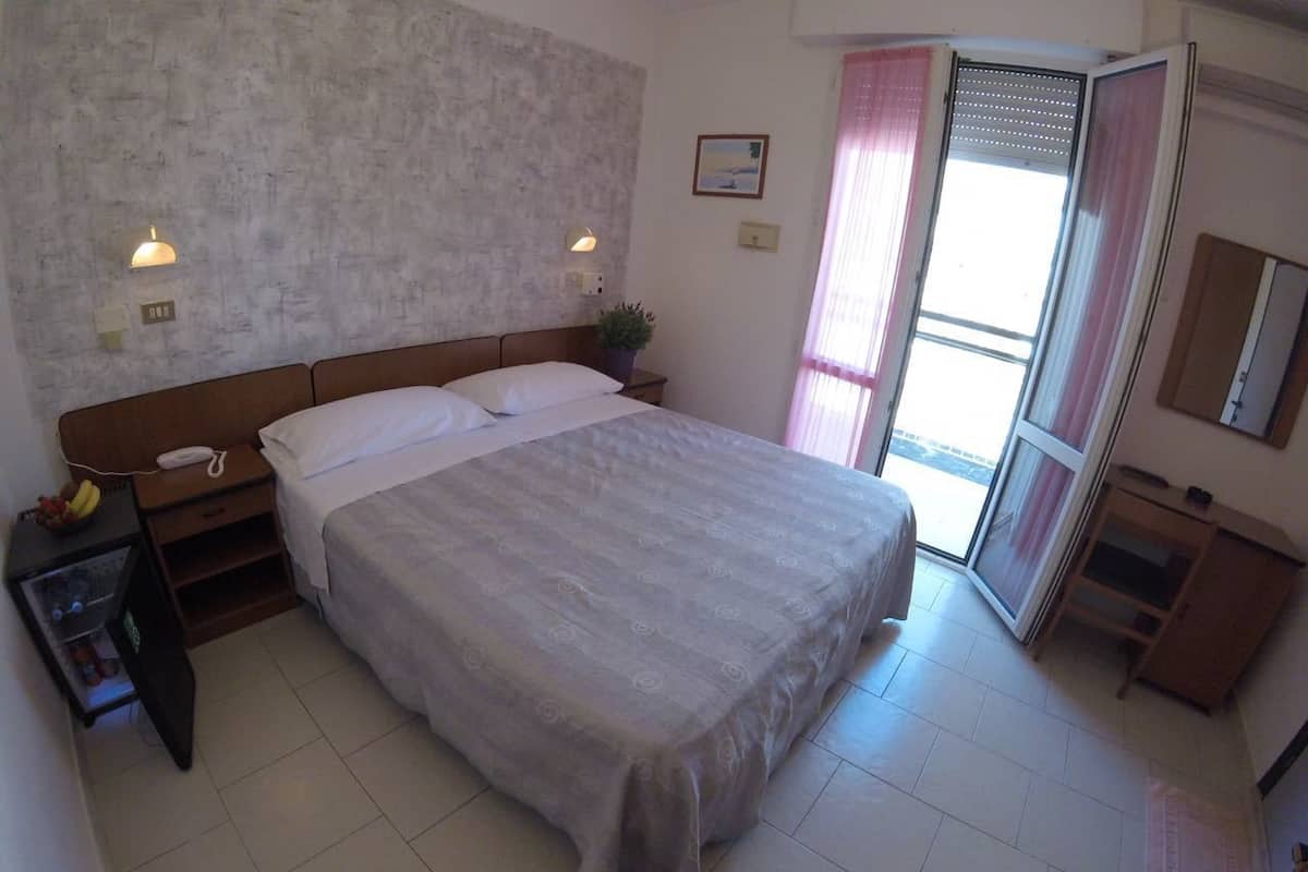 A bedroom with a large bed, a bedside table with a lamp, a small plant, and a window with curtains.