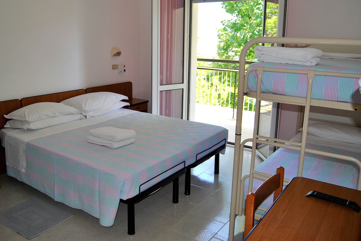 A bedroom with a single bed, a wooden nightstand, a table with a remote control, and a bunk bed with towels on top.
