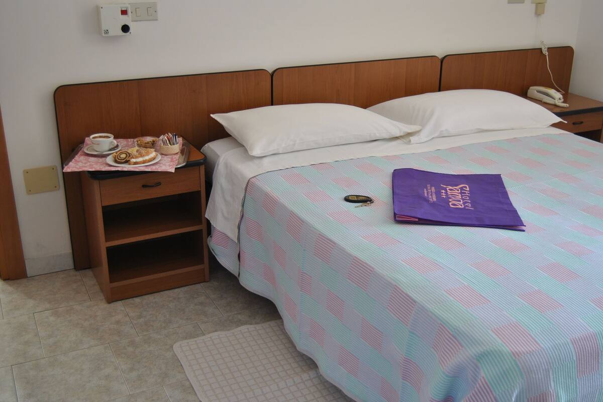 A neatly made bed with a patterned bedspread, a bedside table with a cup of coffee and pastries, and a purple towel on the bed.