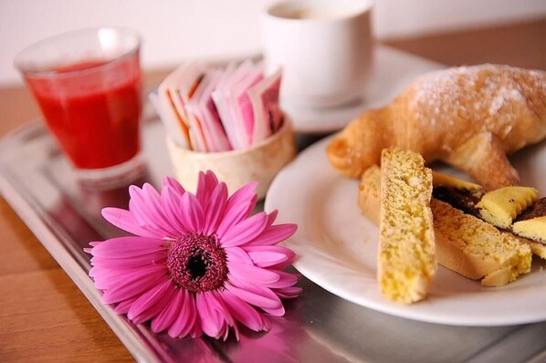 A tray with a pink flower, a glass of red drink, a container of pink and white items, a white cup, and a plate with a croissant and bread slices.