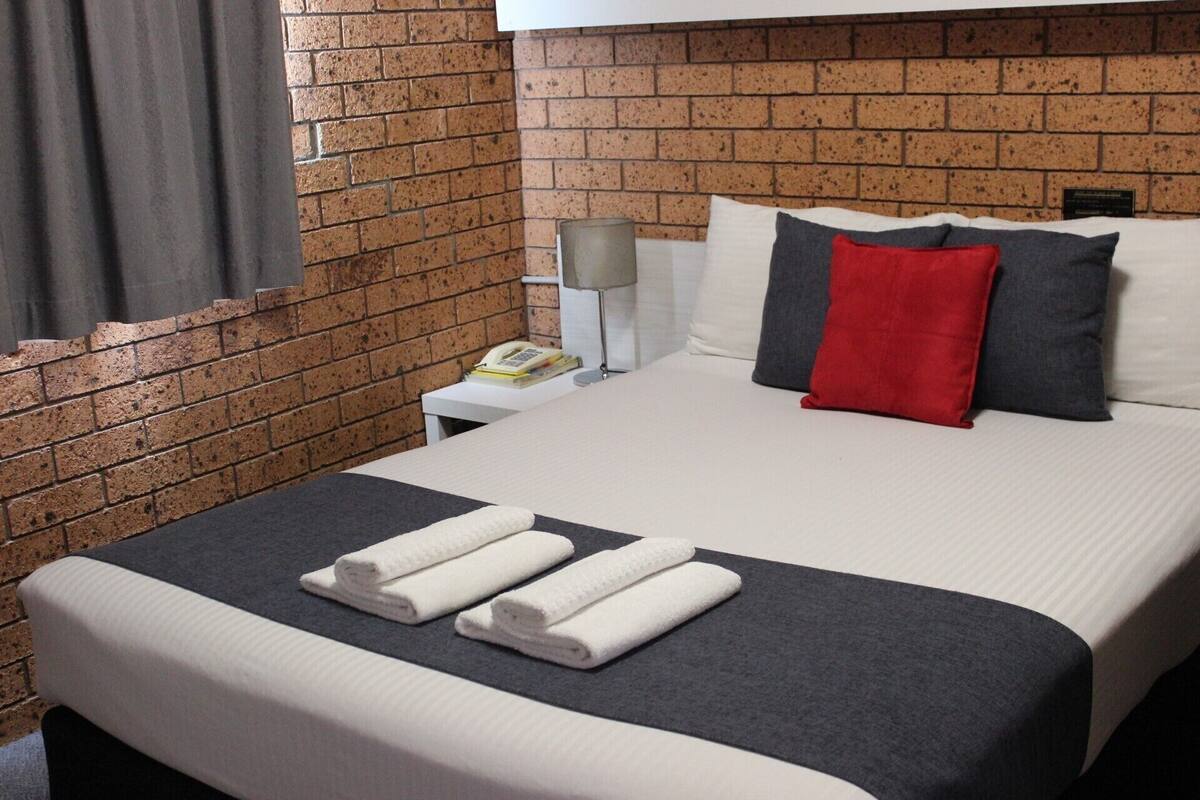 A hotel room with a brick wall, a bed with white and grey bedding, a red pillow, and a folded white towel on the bed.
