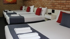 A hotel room with four beds, each with white linens and red and grey pillows. There is a brick wall and a painting above the beds. - Golden Harvest Motor Inn (Moree)