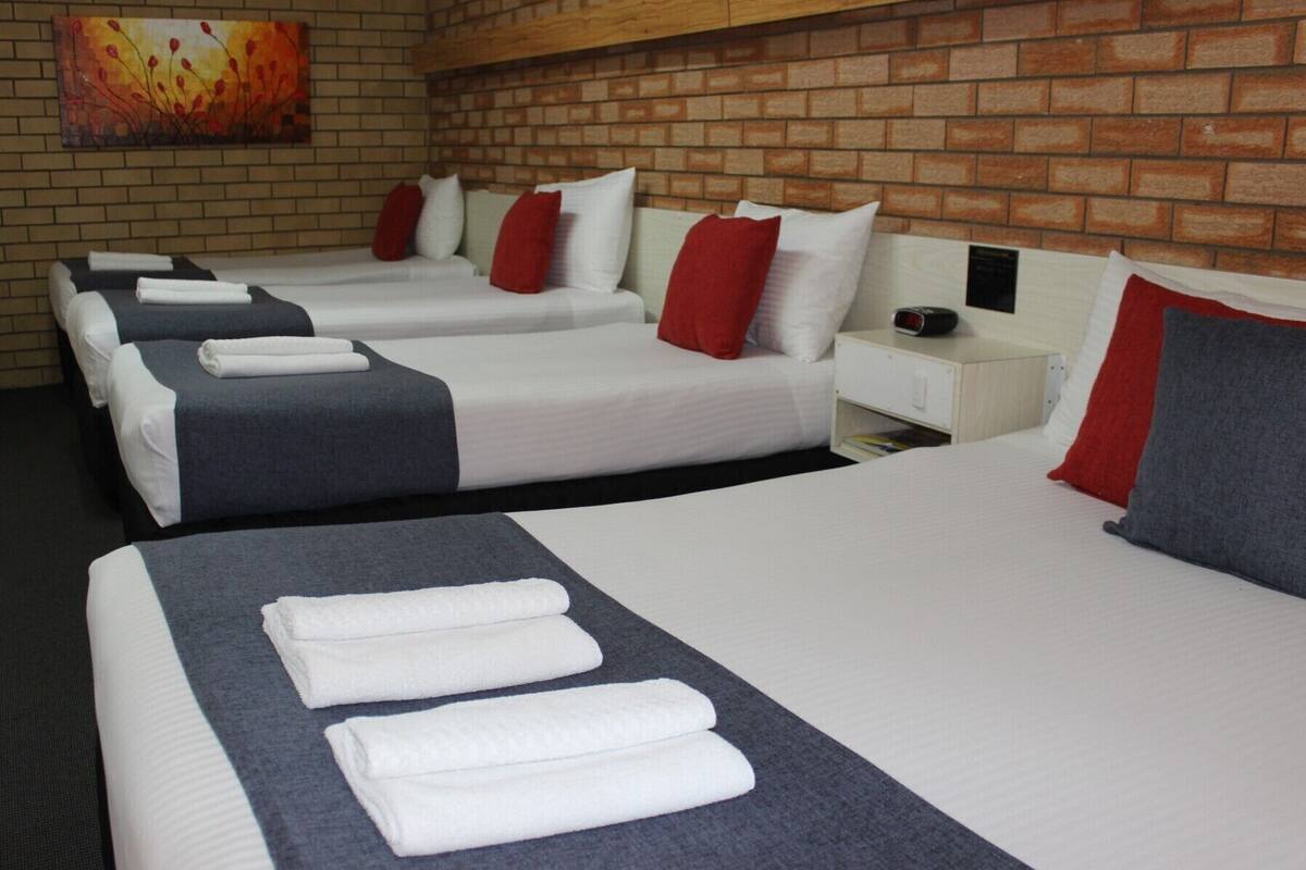 A hotel room with four beds, each with white linens and red and grey pillows. There is a brick wall and a painting above the beds.