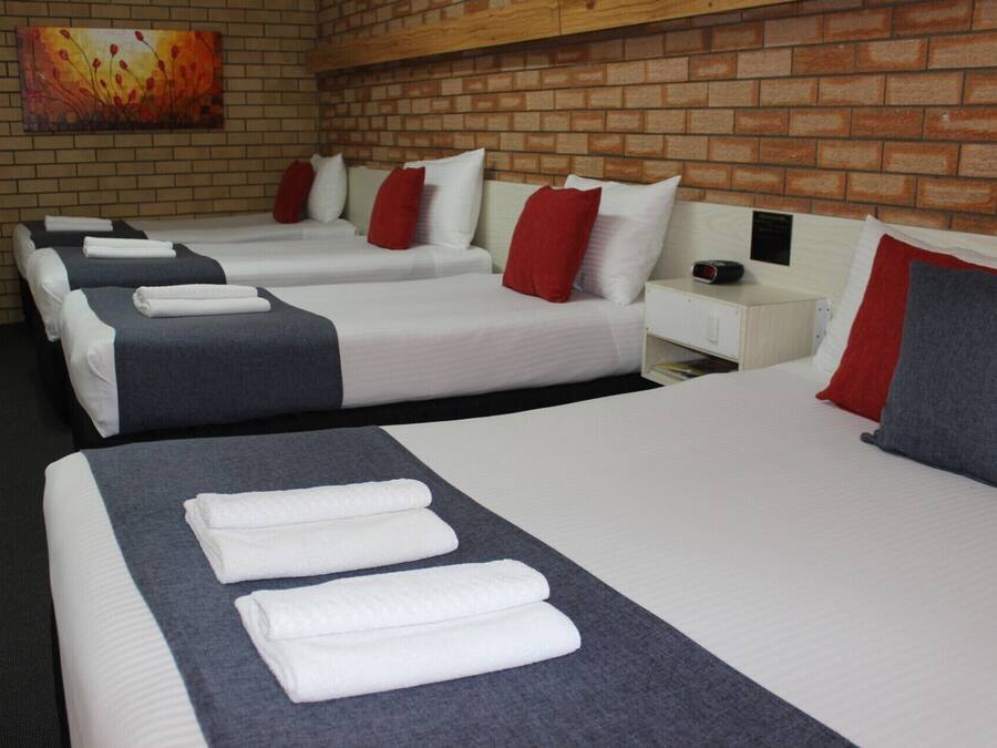 Quarto de hotel com quatro camas, cada uma com lençóis brancos e almofadas vermelhas e cinzentas. Há uma parede de tijolos e uma pintura acima das camas.