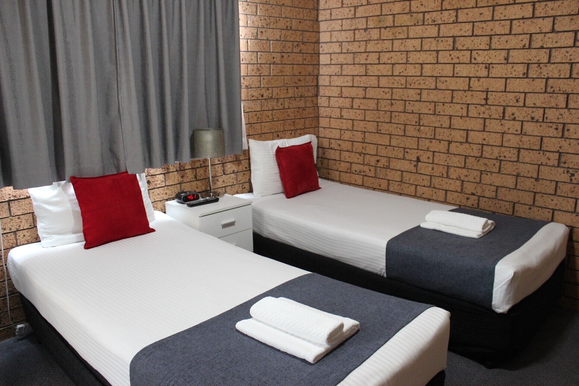 A hotel room with two beds, grey curtains, a brick wall, and a nightstand with a lamp.