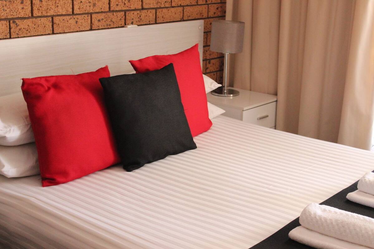 A neatly made bed with striped bedding, two pillows (one red, one black), a bedside lamp, and a nightstand.