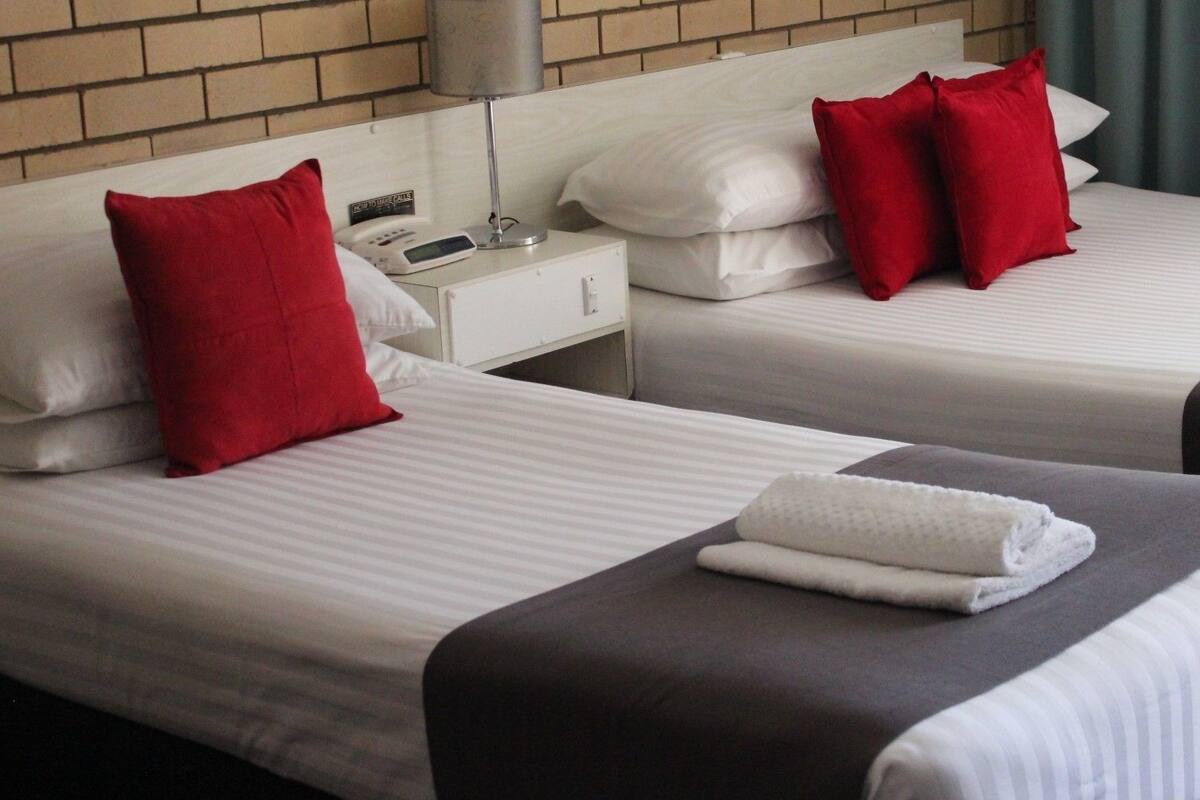 Two single beds with white linens and red pillows, a bedside table with a phone and a lamp, and a folded towel on one bed.