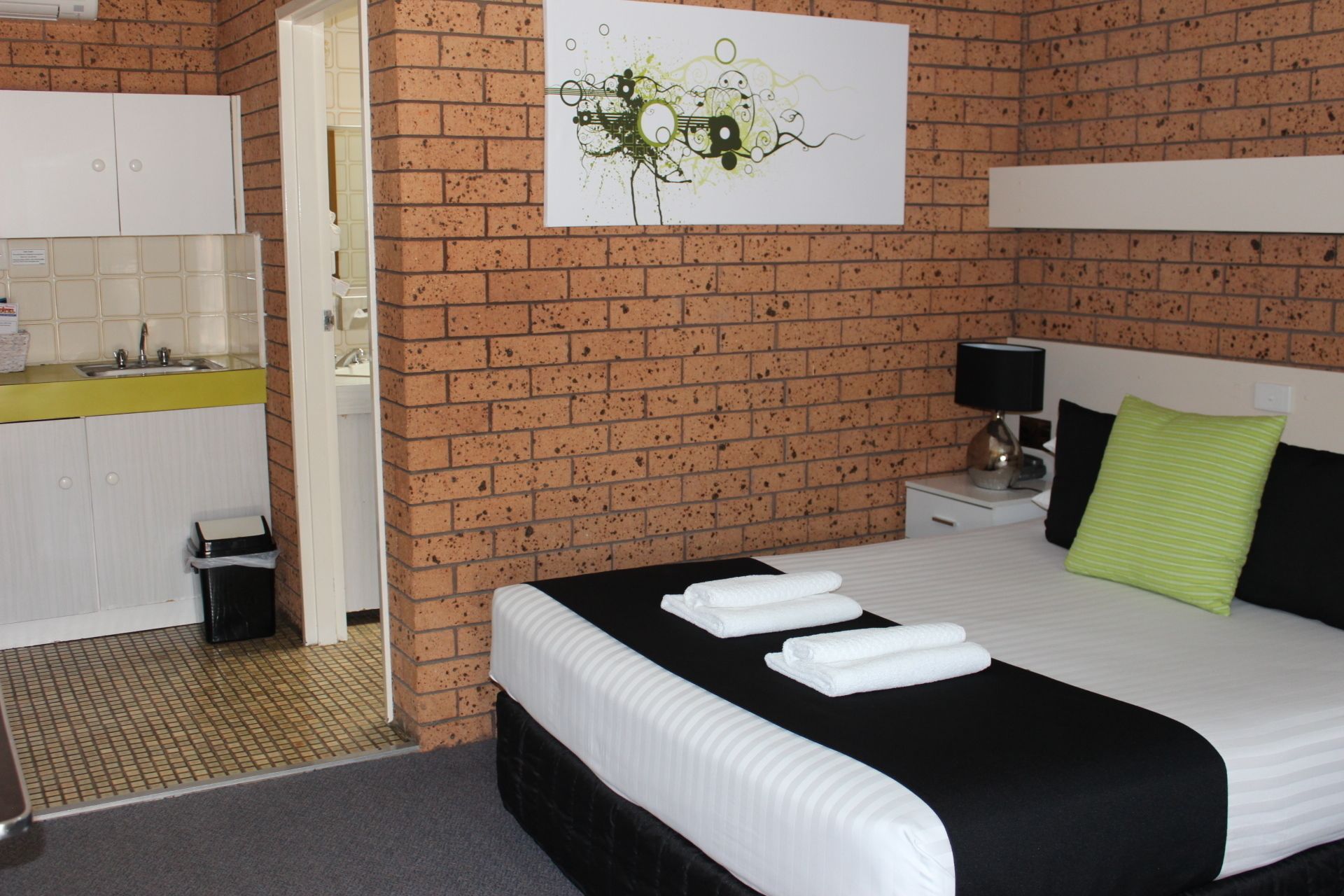 Ein Hotelzimmer mit Backsteinwand, einem Bett mit weißer Bettwäsche, einem Nachttisch mit Lampe und einer kleinen Küchenzeile.