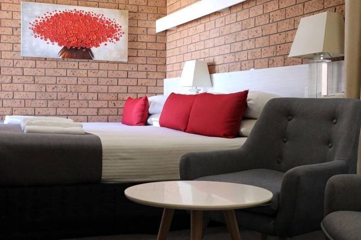 A hotel room with a brick wall, a bed with red and white bedding, a grey armchair, a round table, and a painting of red flowers.