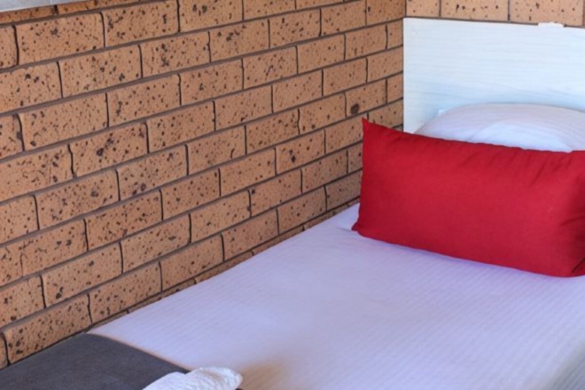 A single bed with a white mattress and a red pillow against a brick wall.
