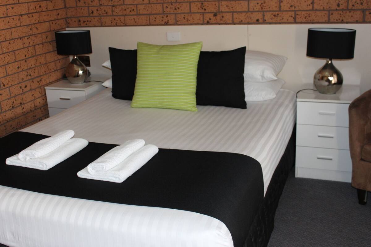 A neatly made bed with white linens and two towels, a green striped pillow, and black pillows on a white headboard. There are two bedside tables with lamps, a brown chair, and a white dresser.