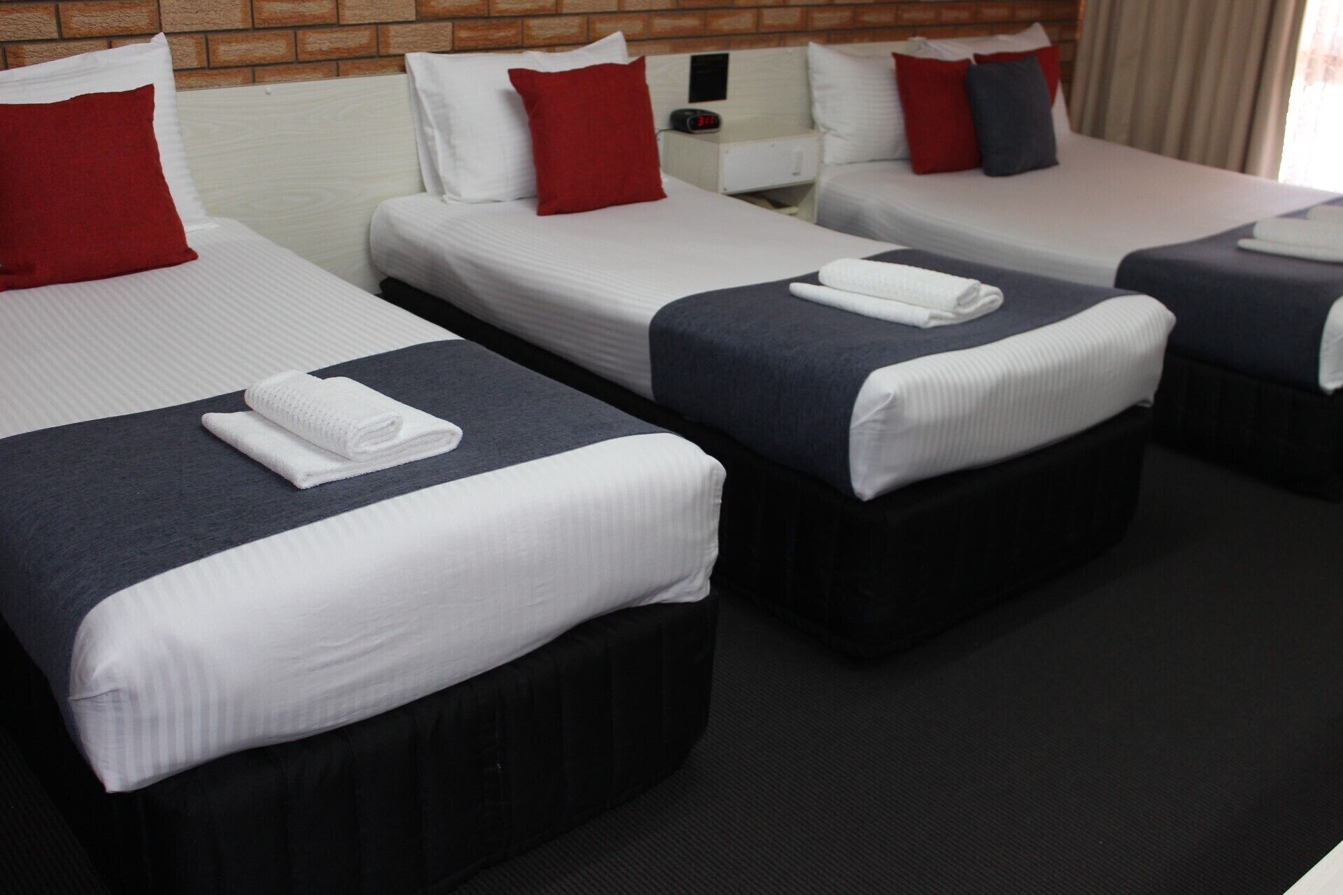 A hotel room with three single beds, each with a white mattress, dark headboard, and a red pillow.