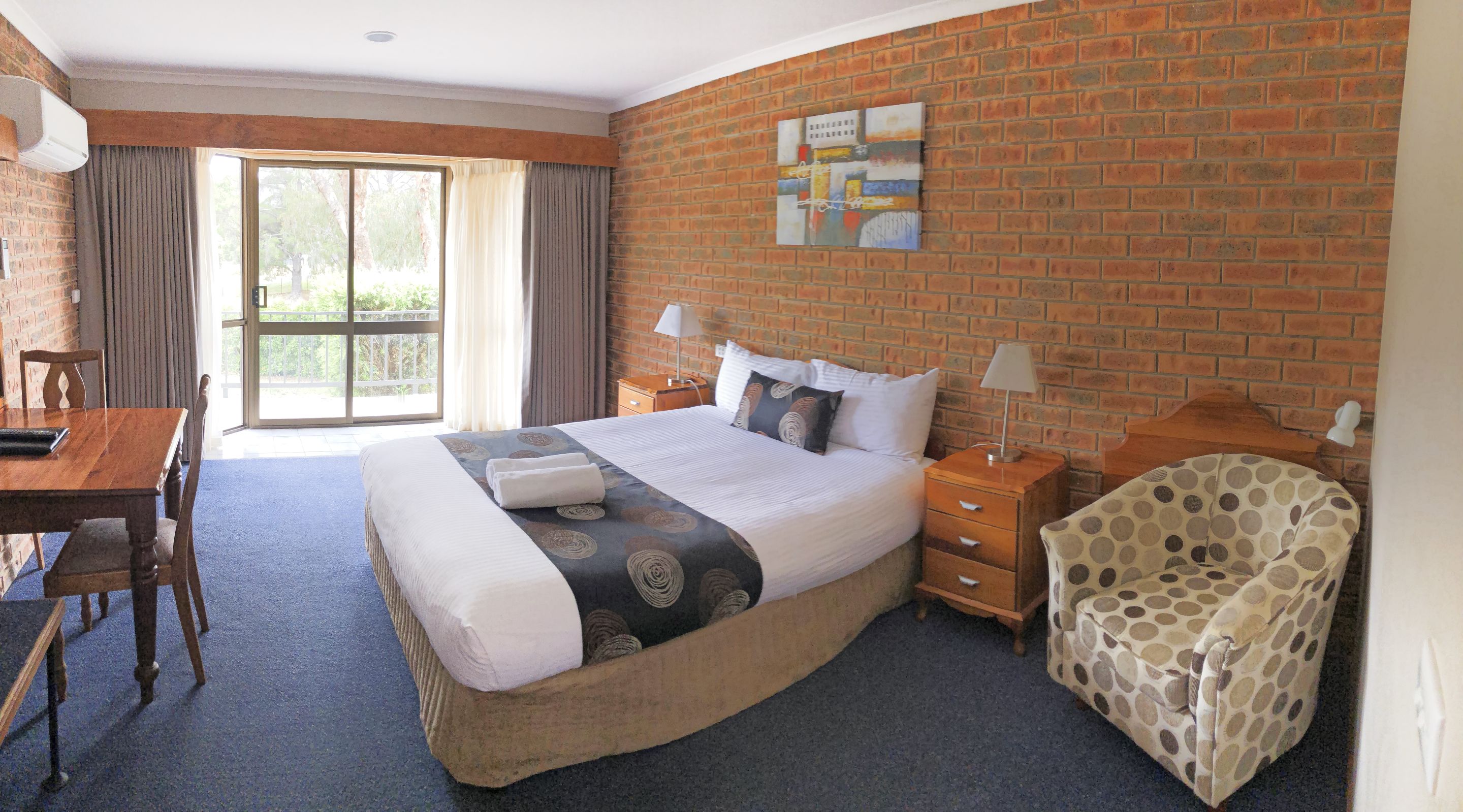 Quarto de hotel com cama grande, escrivaninha, cadeira e vista por uma porta de vidro.