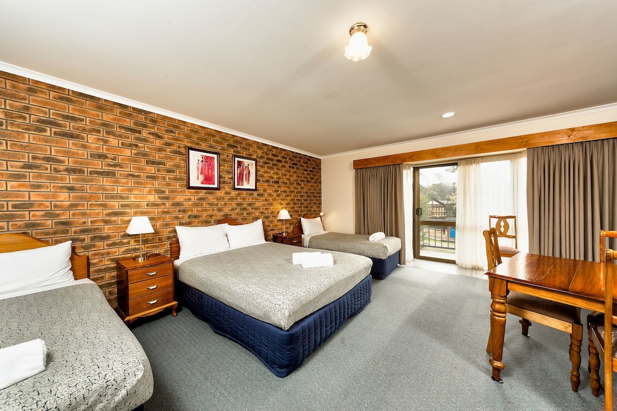 A hotel room with two beds, a wooden dining table, a brick wall, and a balcony with curtains.