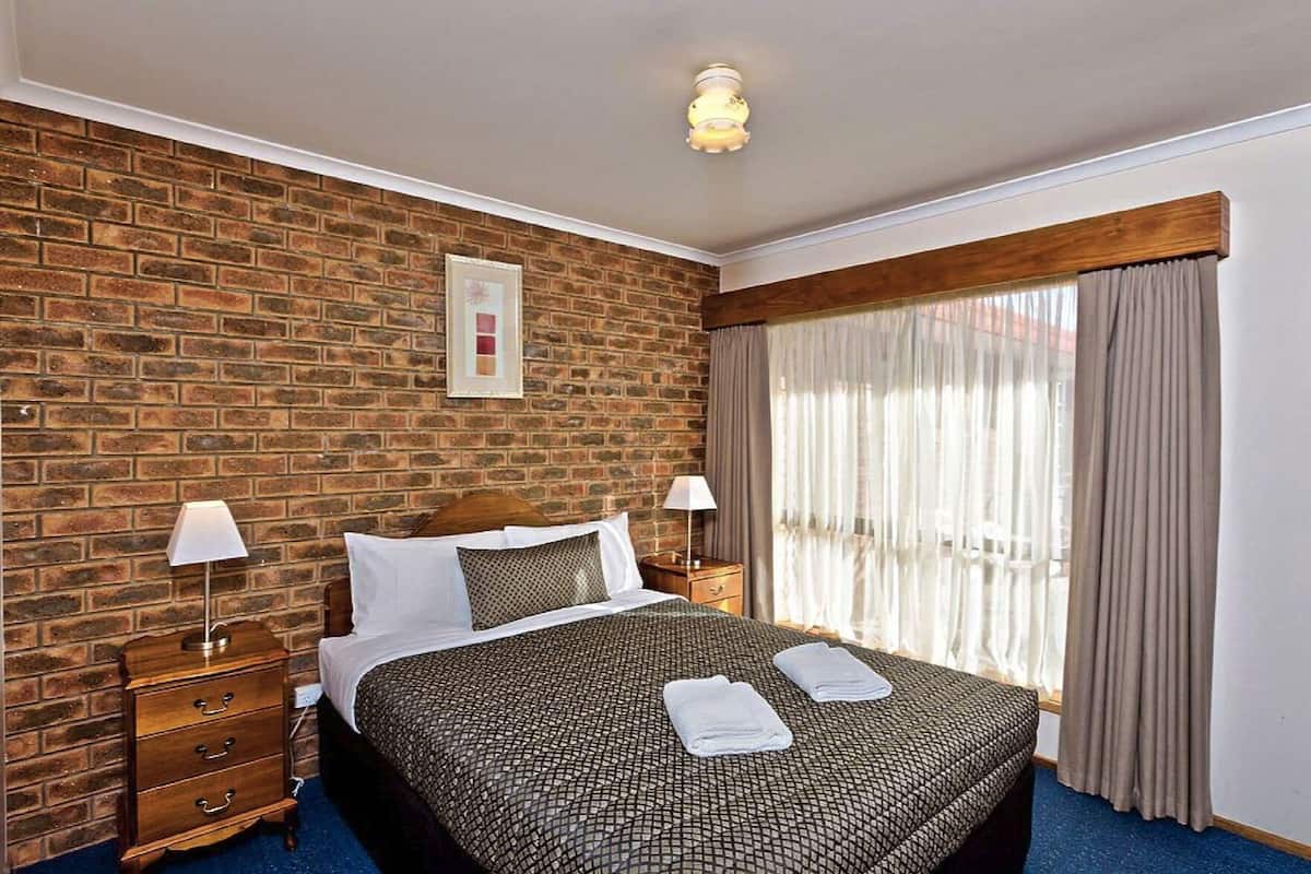 A hotel room with a brick wall, a bed with a patterned bedspread, two bedside tables with lamps, and a window with curtains.