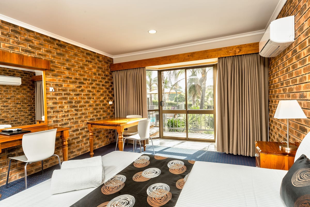 A hotel room with a brick wall, a desk with a chair, a bed with a patterned bedspread, and a large window with curtains.