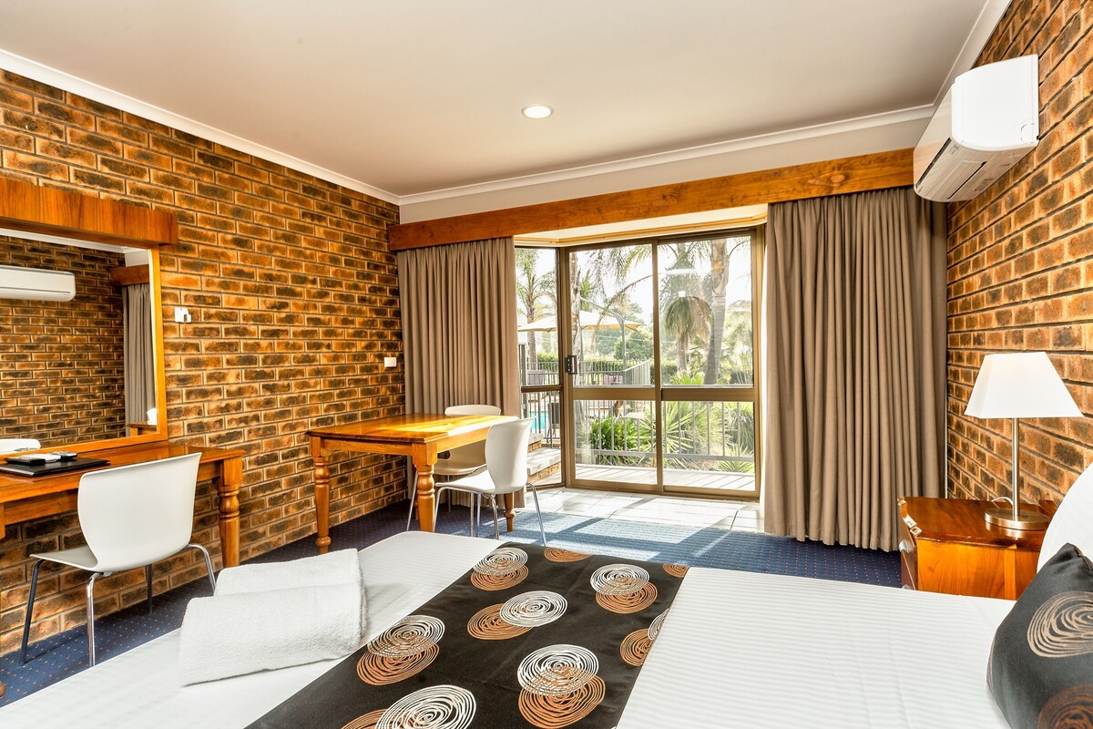 A hotel room with a brick wall, a desk with a chair, a bed with a patterned bedspread, and a large window with curtains.