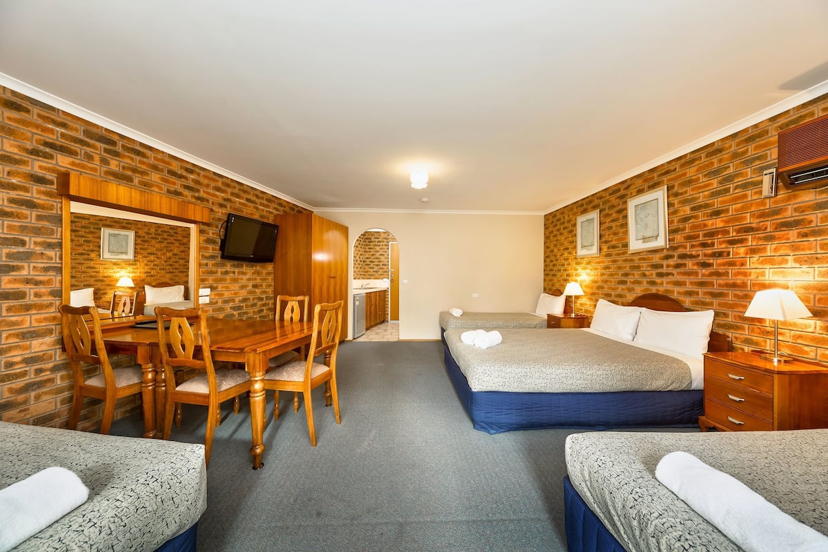 A hotel room with a brick wall, a wooden dining table with chairs, two beds with pillows, a TV mounted on the wall, and a small kitchen area.