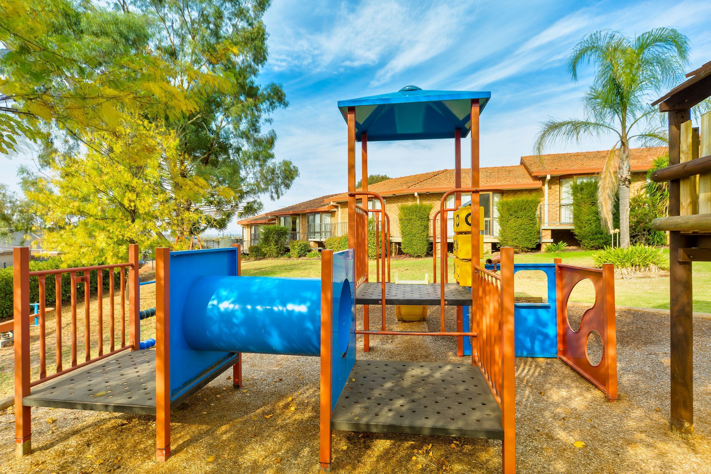 A playground with a blue slide and climbing structures, surrounded by trees and a building in the background.