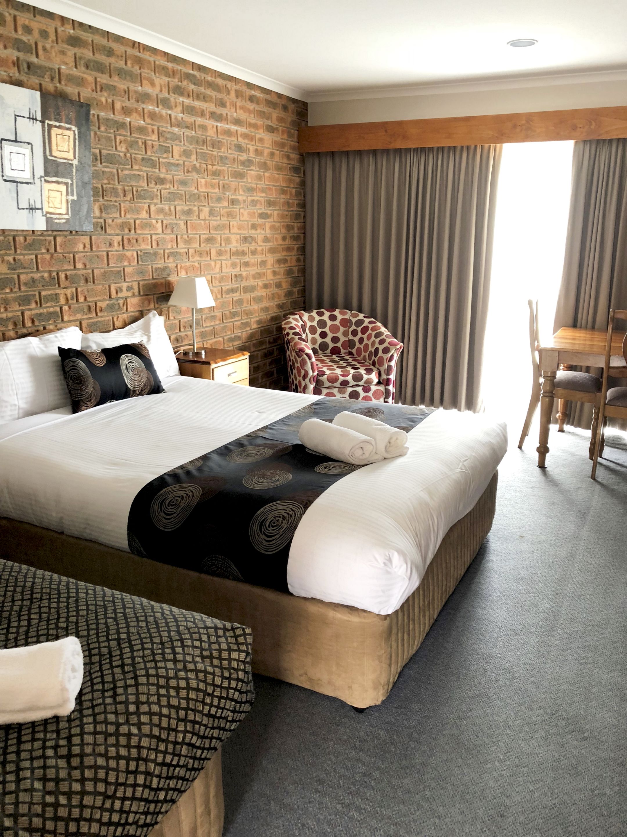 A hotel room with a large bed, a brick wall, a window with curtains, a chair, and a table.