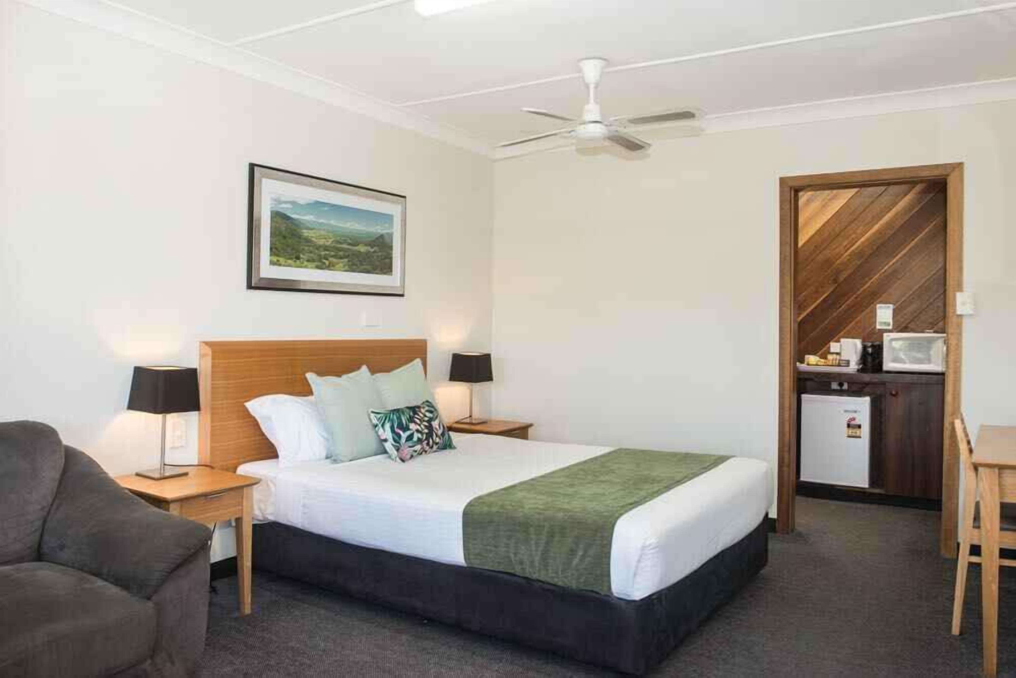 A hotel room with a bed, a desk, a chair, a small kitchenette, and a ceiling fan.