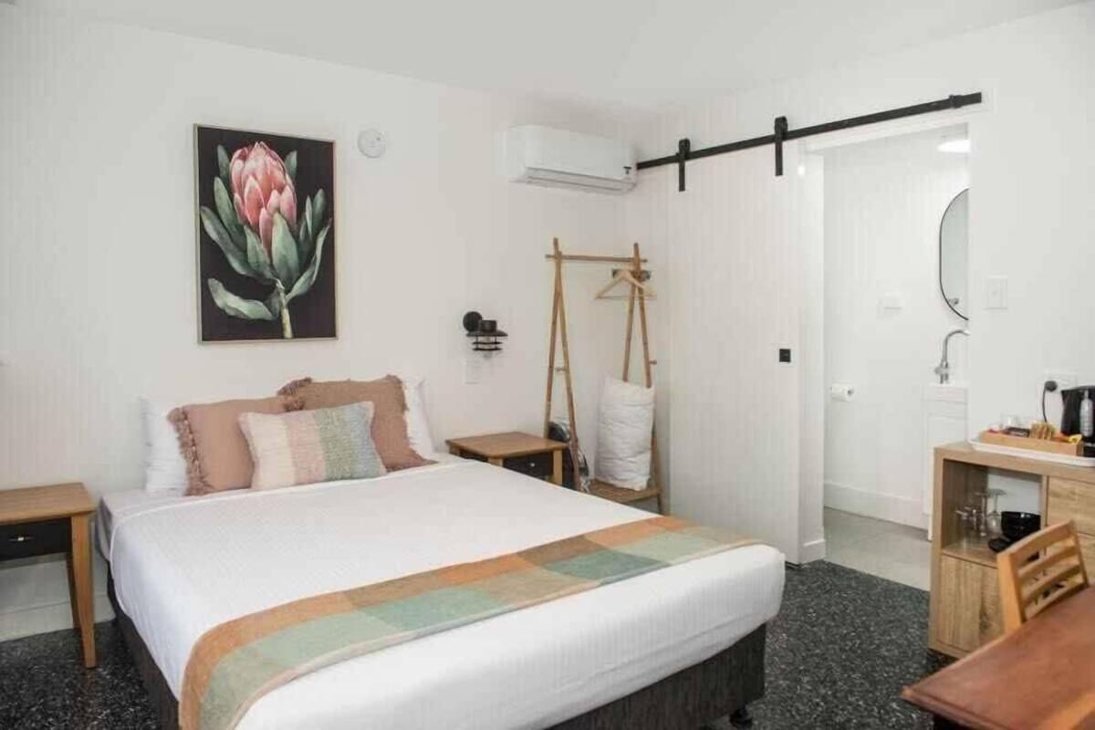 A bedroom with a bed, a bedside table, a hanging light, a wardrobe, a mirror, and a painting of a flower.