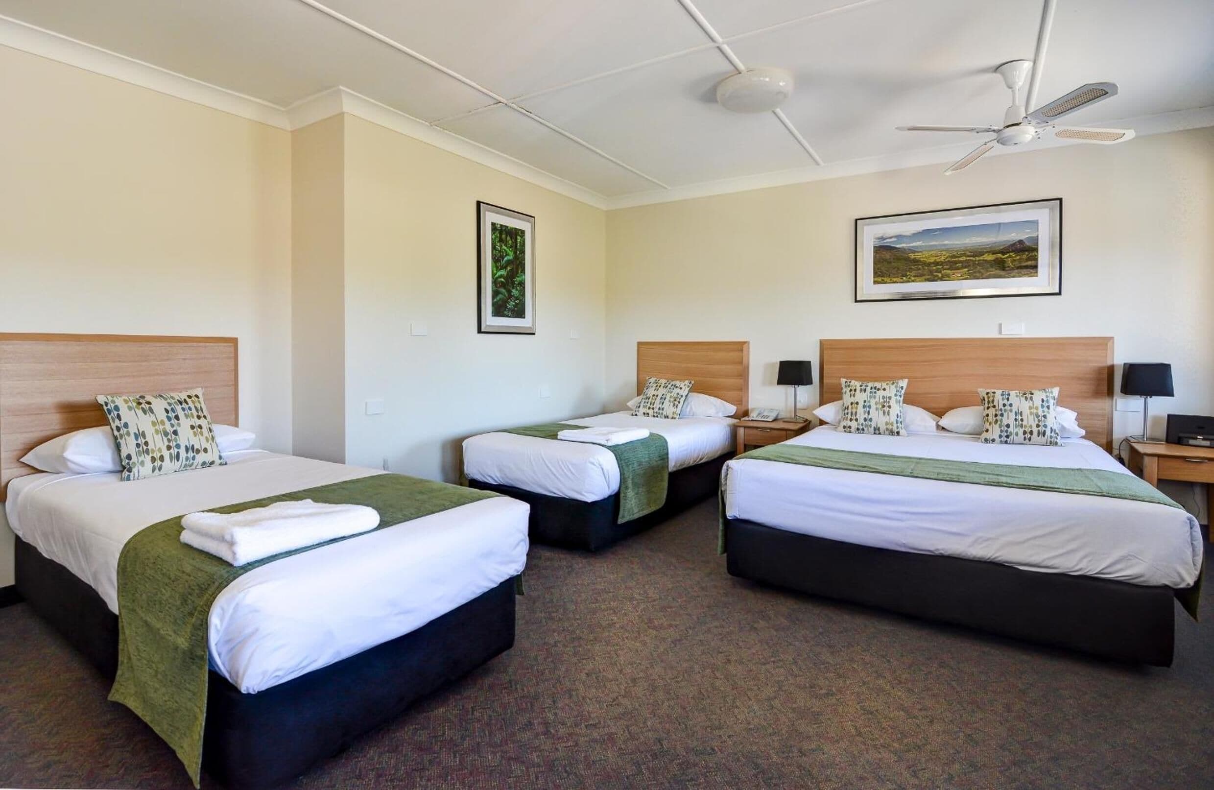 A hotel room with three single beds, a ceiling fan, a framed landscape picture, and a small table with a lamp.