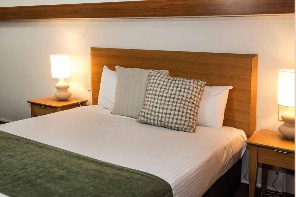 A neatly made bed with a wooden headboard, white bedding, and a green throw blanket. There are two bedside tables with lamps, one on each side of the bed.