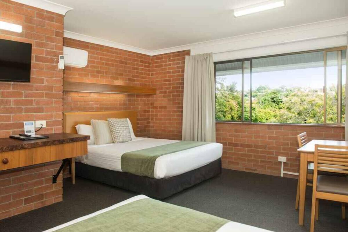 A hotel room with a brick wall, a bed with a green blanket, a desk with a chair, and a window with a view of greenery.