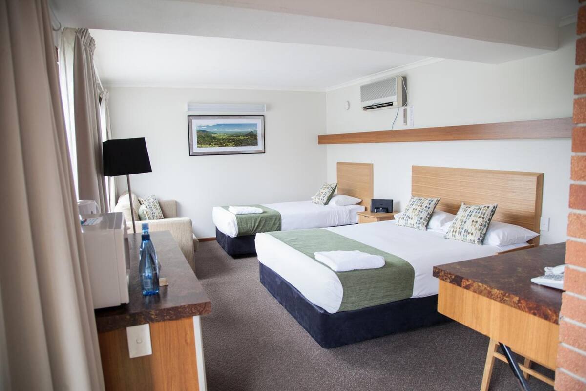 A hotel room with two beds, a desk, a chair, a lamp, a picture on the wall, and an air conditioning unit.