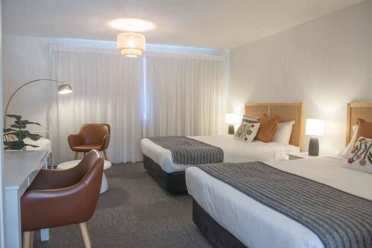 A hotel room with two beds, a desk, a chair, and a lamp.