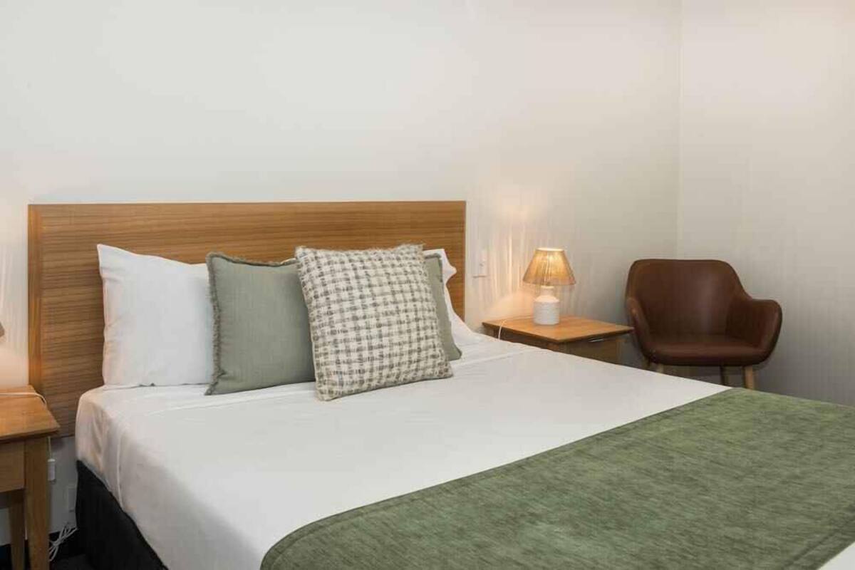 A neatly made bed with a green throw blanket, a wooden headboard, and two pillows. There is a brown leather chair and a bedside table with a lamp.
