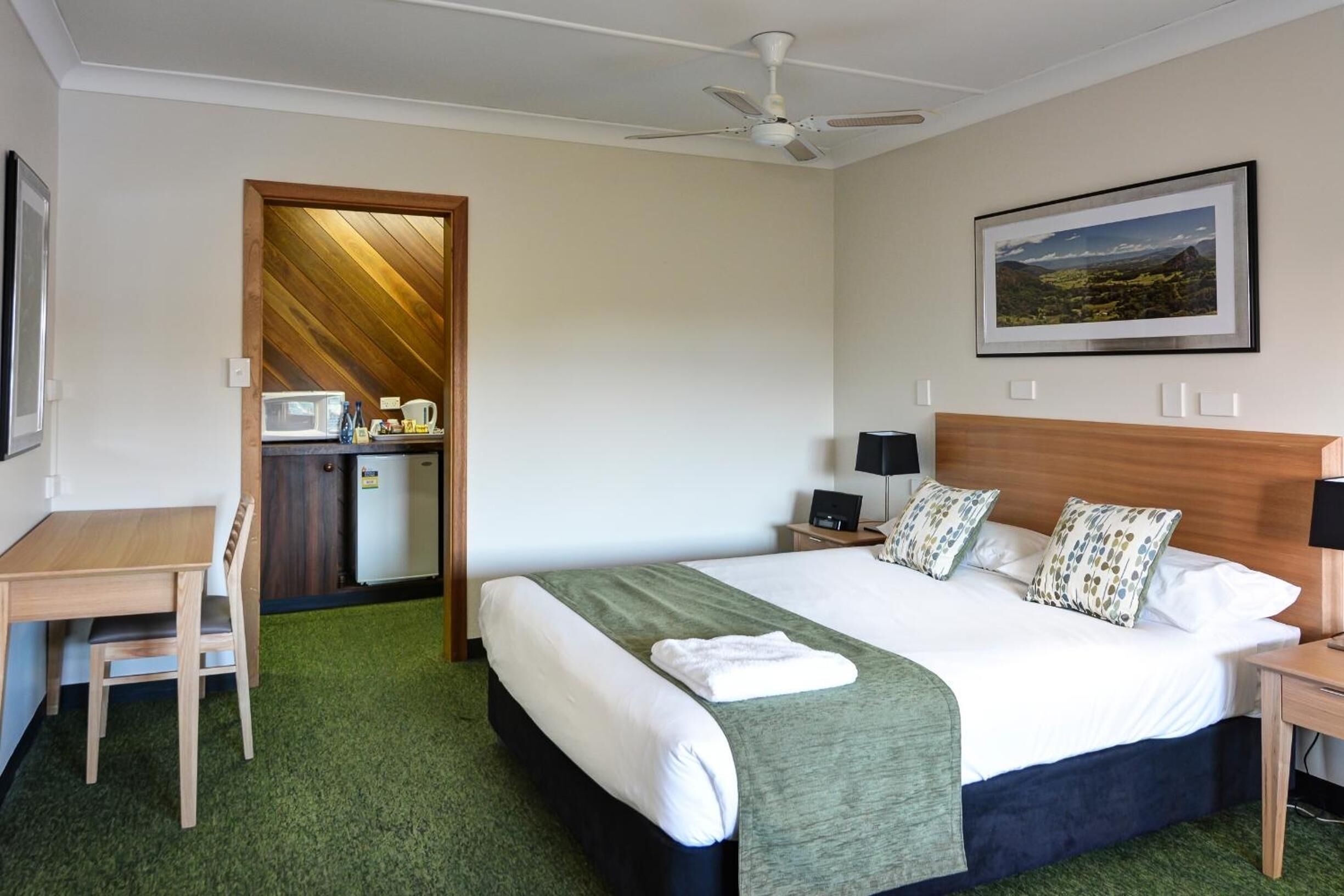 A hotel room with a bed, a desk, a chair, a microwave, a refrigerator, a ceiling fan, and a framed picture on the wall.