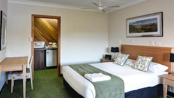 A hotel room with a bed, a desk, a chair, a microwave, a refrigerator, a ceiling fan, and a framed picture on the wall.