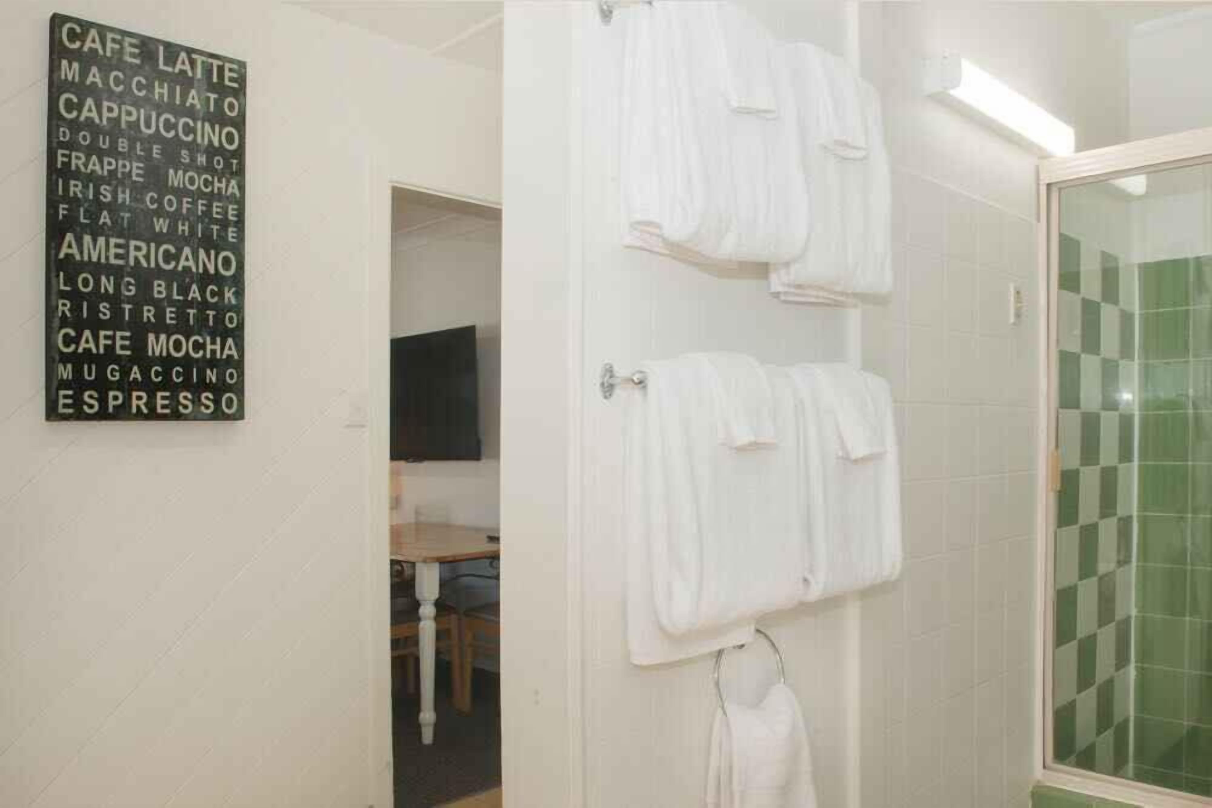 A bathroom with white tiles, a shower with a green glass door, and a wall-mounted sign listing coffee and espresso types.