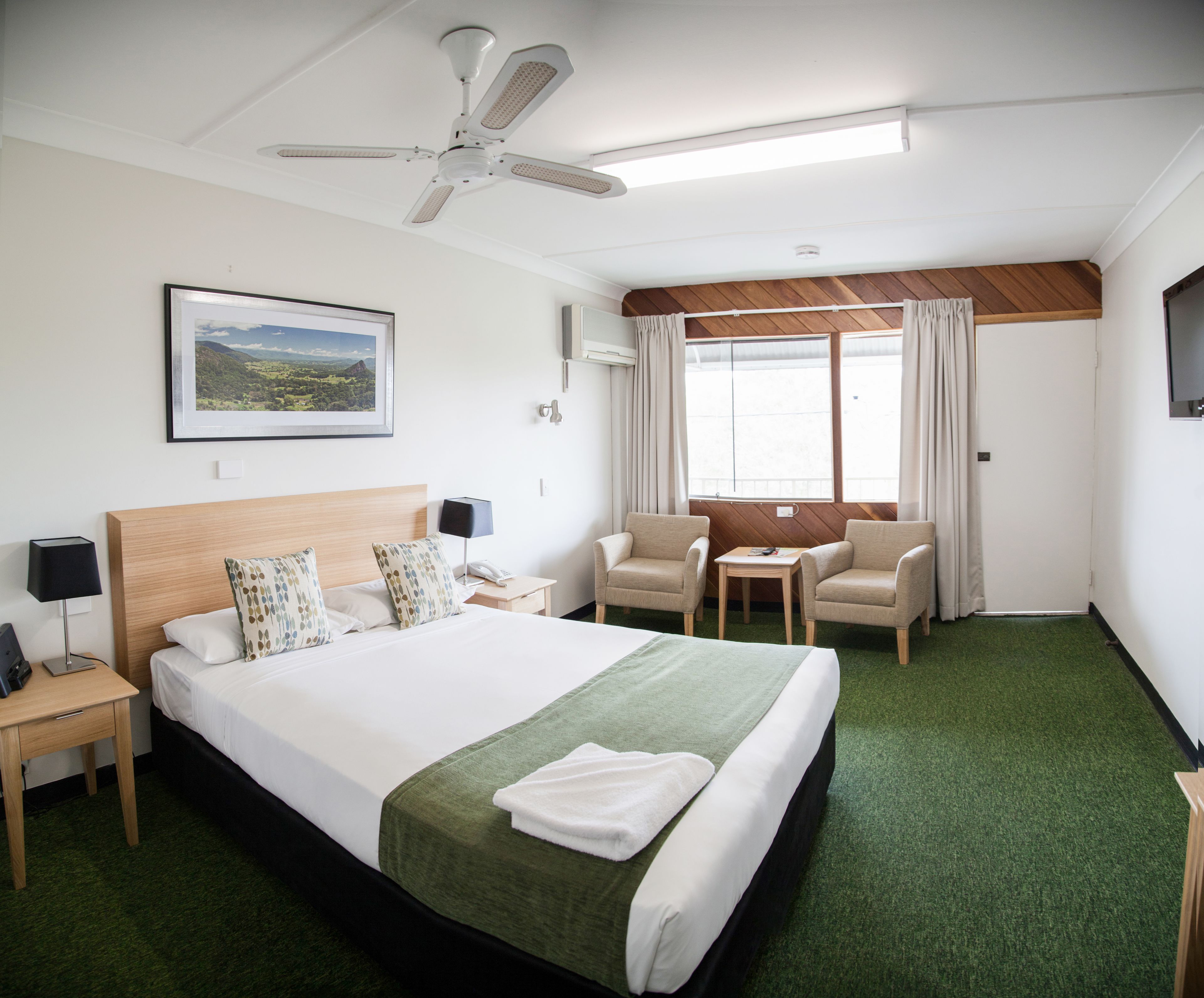 A hotel room with a large bed, a desk, two chairs, a ceiling fan, a window with curtains, and a framed picture on the wall.