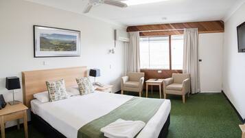 A hotel room with a large bed, a desk, two chairs, a ceiling fan, a window with curtains, and a framed picture on the wall.