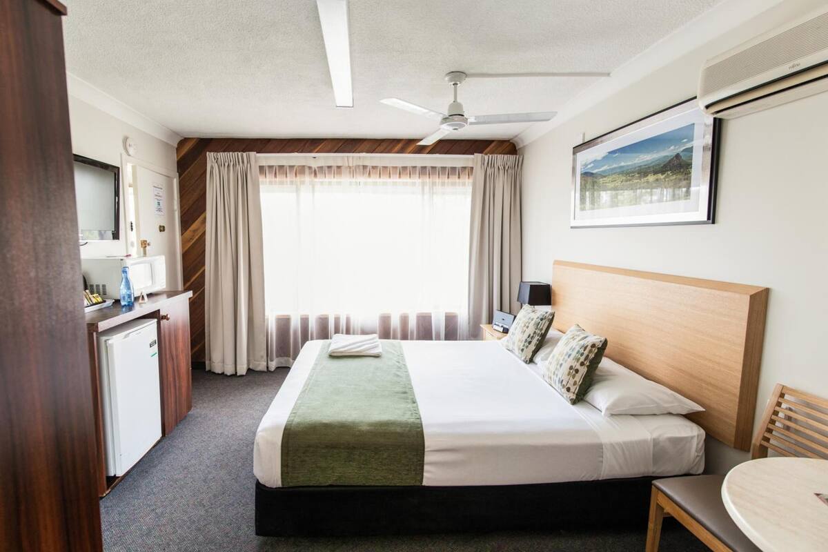 A hotel room with a bed, a desk, a chair, a table, a ceiling fan, and a window with curtains.
